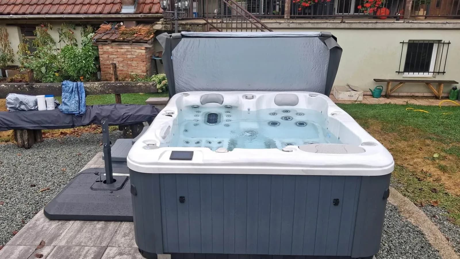 Spa installé dans le jardin d'une maison, sur une dalle au bord de la pelouse, couverture relevée