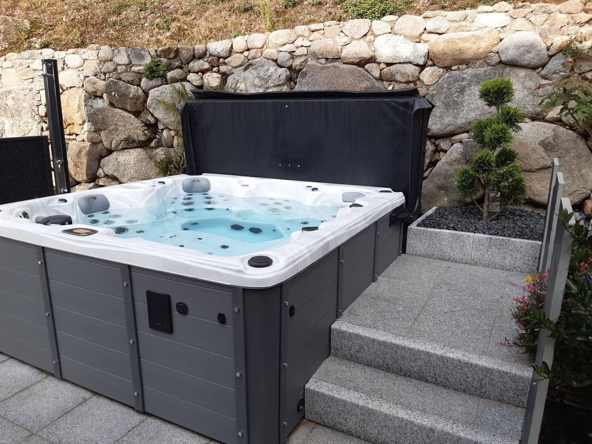 Spa carré à coque acrylique blanche et tablier gris installé sur une terrasse, couverture relevée et eau bleutée